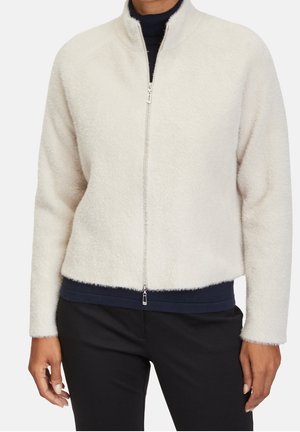 Osoba ubrana w kremową polarową kurtkę zapinaną na zamek, na granatową koszulę i czarne spodnie, stojąca na białym tle.