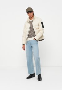 Doudoune beige avec un accent noir sur la manche, superposée à un pull à motifs, associée à un jean bleu clair et des bottines noires.
