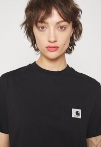 T-shirt en coton noir avec un col rond, des manches courtes et une poche sur la poitrine gauche avec un patch logo contrasté.