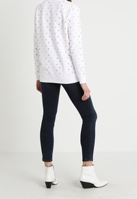 Persona che indossa un top bianco a maniche lunghe con motivo, jeans slim scuri e stivali con tacco bianchi alla caviglia, in piedi contro uno sfondo chiaro e semplice.