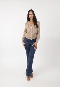 Beige geribde cardigan met gouden knopen en twee borstzakken, gecombineerd met blauwe uitlopende jeans en sandalen met doorzichtige hakken op een witte achtergrond.