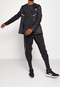 Man i svart Adidas träningsjacka och byxor, samt svarta och vita Adidas sneakers, står med en arm sträckt ut.