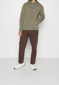 Osoba ubrana w oliwkowozieloną bluzę z kapturem, brązowe spodnie i białe trampki, stojąca z jedną ręką w kieszeni na jednolitym tle.