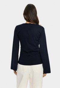 Femme aux longs cheveux bruns de dos, portant un haut à manches longues bleu marine et un pantalon écru devant un fond uni.