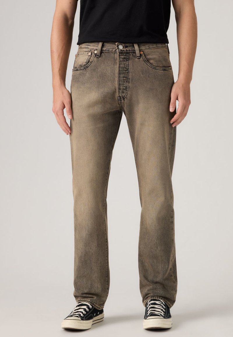 Άνδρας που φοράει ξεθωριασμένα καφέ τζιν παντελόνια με ευθείες μπότες και μαύρα χαμηλά αθλητικά παπούτσια, στεκόμενος μπροστά σε απλό φόντο.