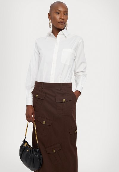 Weißes Button-Up-Hemd mit einer Tasche, kombiniert mit einem braunen Rock mit mehreren aufgesetzten Taschen und goldenen Knöpfen, gehalten mit einer schwarzen Handtasche.