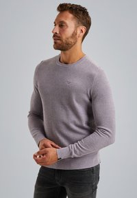 Vijolična pletena pulover s krožno ovratnikom in rebrastimi manšetami. Nosijo ga z temnimi kavbojkami, ki imajo majhen logotip na prsih.
