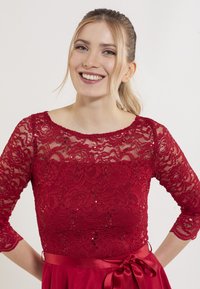 Robe rouge avec un corsage en dentelle orné de motifs floraux et de sequins, manches trois-quarts et ceinture en satin nouée à la taille.
