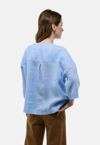 Blouse en lin bleu clair au fit ample, avec des manches trois-quarts et un détail de pli au dos. Portée avec un pantalon taille haute marron.