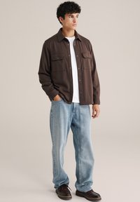 Barna, hosszú ujjú ing gombos elülső résszel és mellzsebekkel, világoskék egyenes szárú farmernadrággal és sötét cipővel párosítva. Göndör haj látható.