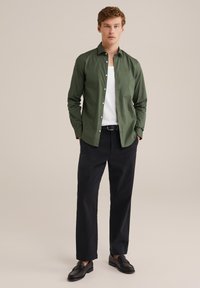 Hombre de pie con una camisa verde de botones abierta sobre una camiseta blanca, pantalones negros con cinturón y mocasines negros, sobre un fondo beige liso.