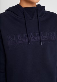 Sudadera azul marino con un parche de logo texturizado, capucha con cordón y cuello acanalado. Presenta un ajuste relajado y una textura de tela suave.