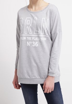Person iført en lysegrå sweatshirt med "BRLN Berlin the place to be No 36" trykt i hvid, kombineret med mørkeblå jeans.