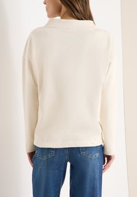 Cremefarbenes Langarmshirt mit Kragen, glatter Textur und geradem Saum, kombiniert mit blauen Jeans mit Gesäßtaschen.