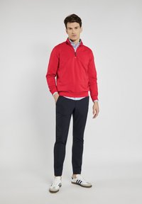 Czerwony sweter z kołnierzem na pół zamek, w zestawieniu z ciemnoniebieskimi spodniami. Model nosi białe trampki w czarne paski, pod którymi widoczna jest niebieska w paski koszula.