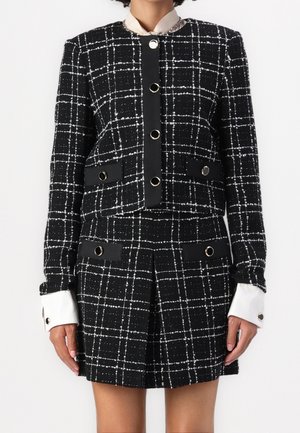 Donna che indossa un blazer a quadri bianco e nero e una minigonna plissettata abbinata con grandi bottoni e polsini della camicia bianchi visibili.