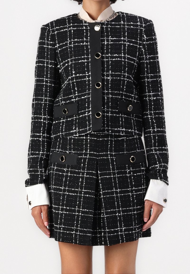 Femme portant un blazer à carreaux noir et blanc et une mini-jupe plissée assortie avec de gros boutons et des poignets de chemise blancs visibles.