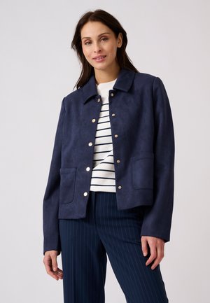 Mujer con chaqueta azul marino abotonada sobre una camiseta blanca con rayas y pantalones azul marino de rayas finas, de pie frente a un fondo liso y claro.