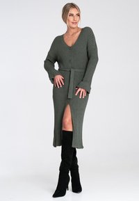 Olivengrünes, geripptes Midi-Kleid mit V-Ausschnitt, Knopfdetails, tailliertem Gürtel und einem oberschenkelhohen Schlitz, kombiniert mit schwarzen kniehohen Stiefeln.