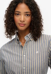 Blå og creme-stribet skjorte med knaplukning og krave, med diskret logo-broderi og klassisk knaplukning foran. Blød tekstur.