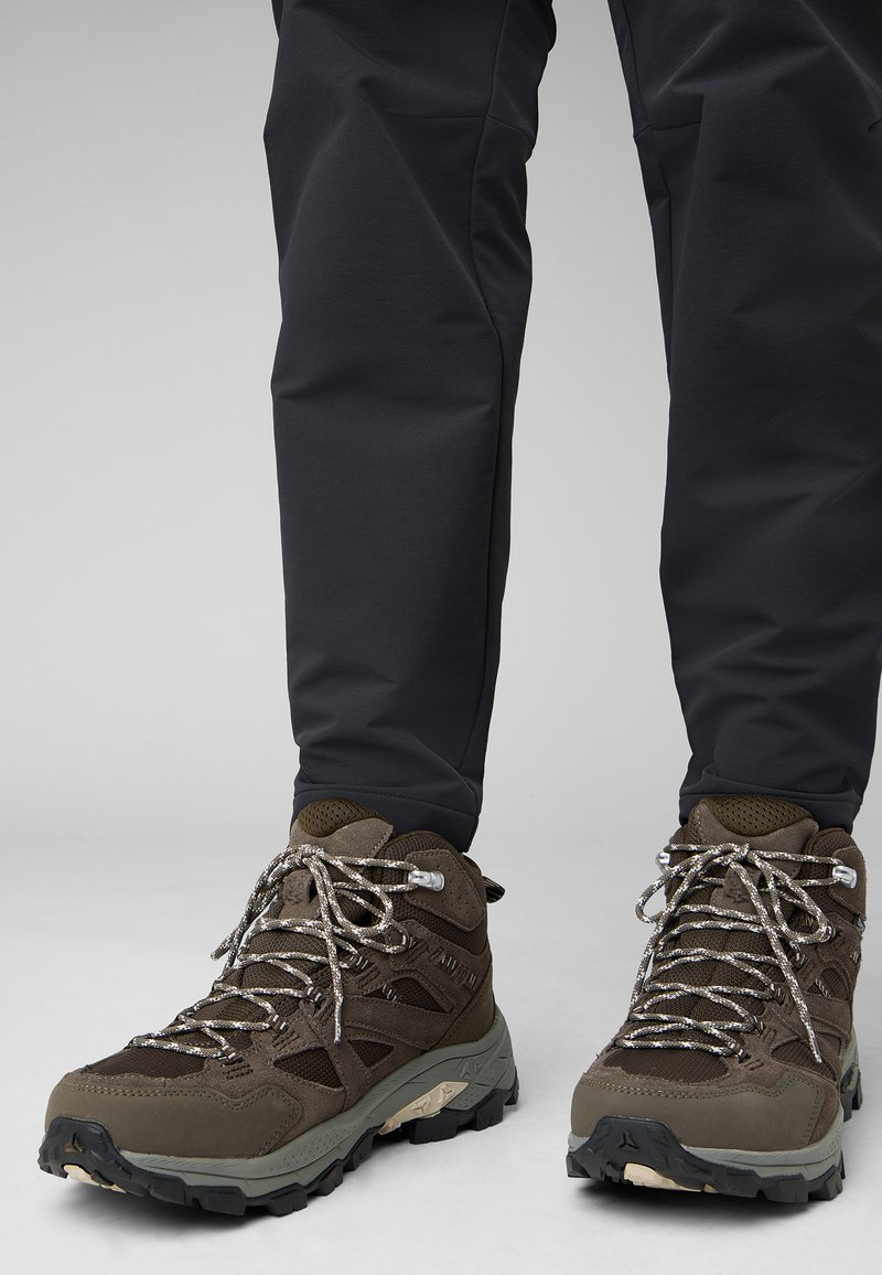 Scarponi da trekking marroni con lacci grigi, pannelli in rete e suole in gomma resistenti. Pantaloni neri aderenti completano il look da outdoor.