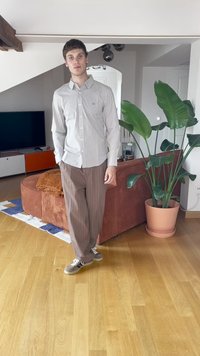 Jovem de camisa às riscas e calças castanhas de pé numa sala de estar com chão de madeira, uma grande planta em vaso e um sofá curvo de cor ferrugem.