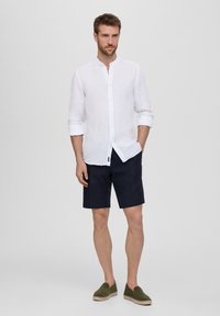 Camicia bianca in lino con colletto alla cinese, pantaloni corti blu navy e espadrillas verdi oliva. Modello in piedi, mani in tasca, sfondo neutro.