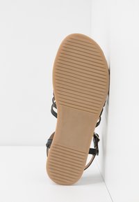Sandale avec semelle en caoutchouc marron clair présentant des rainures horizontales, des brides noires et une boucle métallique sur fond blanc.
