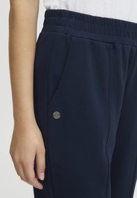 Navyblå träningsbyxor med elastisk midja, med en framficka och en rund metallisk detalj nära fickans kant. Mjuk tygtextur.