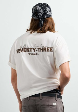 Person mit langen Haaren, die ein schwarzes Bandana mit Muster und ein weißes T-Shirt mit braunem Text "SEVENTY-THREE PEPE JEANS" auf dem Rücken trägt.