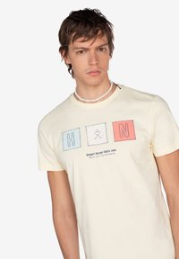 Camiseta de algodón color amarillo claro con un gráfico que presenta las letras H, N y un ícono de calavera en cuadrados coloridos. Diseño de manga corta y cuello redondo.