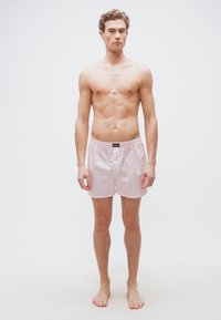 Νεαρός άνδρας με σγουρά μαλλιά, όρθιος και ξυπόλητος, φοράει ροζ ανοιχτόχρωμα και λευκά ριγωτά μποξεράκια, μπροστά σε λευκό φόντο.