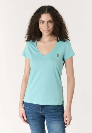 Donna con capelli ricci che indossa una maglietta azzurra a scollo a V e jeans blu, in piedi davanti a uno sfondo chiaro e semplice.