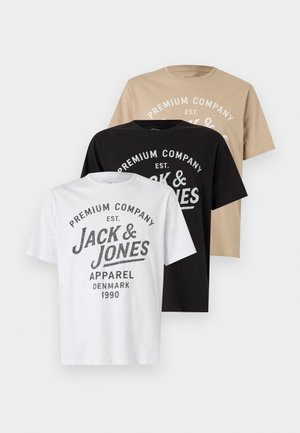 Jeune homme debout, les mains dans les poches, portant un t-shirt blanc imprimé, un short gris, des chaussettes blanches et des baskets blanches à rayures noires.