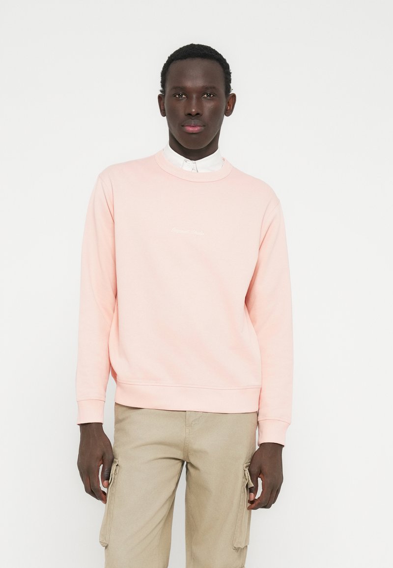 Joven con sudadera rosa claro sobre camisa blanca con cuello y pantalones cargo beige, de pie contra un fondo blanco liso.
