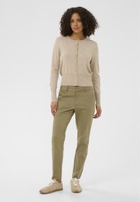 Cardigan beige à boutons, associé à un pantalon vert olive. Le modèle porte des baskets marron clair. La texture lisse et la coupe ajustée améliorent l'apparence.