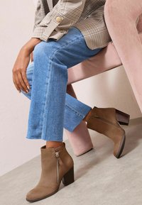 Botas de ante marrón hasta el tobillo con cremallera lateral, tacón grueso y punta redonda, combinadas con pantalones vaqueros de denim azul claro y una camisa de cuadros.