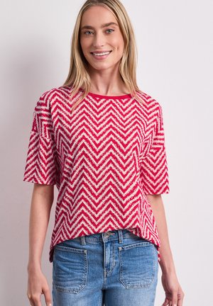 Smilende kvinde iført en rød og hvid zigzag-mønstret top med korte ærmer og blå jeans mod en ensfarvet lys baggrund.