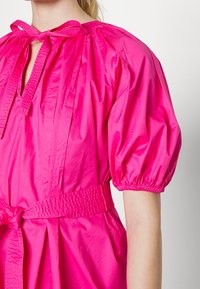 Woman wearing bright pink puff-sleeve dress with a tied bow neckline and a textured belt cinching the waist.