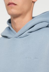 Felpa azzurra chiara realizzata in tessuto morbido, con un discreto logo ricamato "Champion" sul petto e una capiente progettazione del cappuccio.
