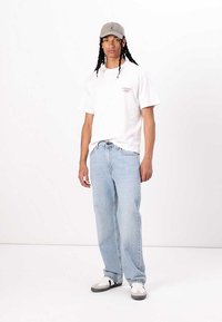 White cotton t-shirt with logo, light blue relaxed-fit jeans, grey cap, and white sneakers with black and grey accents. Simple, casual outfit.
