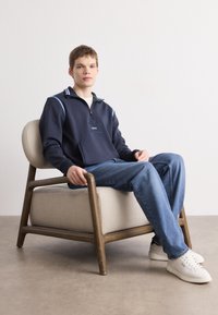 Felpa zip con cappuccio blu navy con accenti blu e verdi, con un logo sul petto. Il modello indossa jeans blu e sneakers bianche, seduto su una sedia chiara.