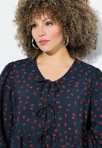 Navy-Kleid mit V-Ausschnitt, Puffärmeln und einem Kordelzugdetail. Verziert mit einem Muster aus kleinen roten Schleifen. Glatte Stofftextur.
