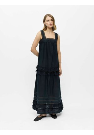 Jeune femme debout en robe longue noire sans manches avec détails en dentelle et chaussures plates noires sur fond blanc uni.