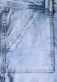 Tissu en denim bleu clair avec une grande poche rectangulaire cousue visible. La texture est lisse avec de légères nuances de décoloration.