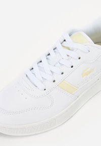 Fehér sportos sneaker sima bőr felsőrésszel, arany díszcsíkkal és textúrált gumitalppal. Laposszemű fűzőkkel és perforált részletezéssel rendelkezik.