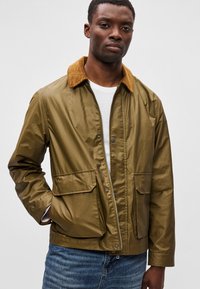 Ελιάς πράσινη ζακέτα με καφέ κορδέλα, που διαθέτει δύο μπροστινές τσέπες, κλείσιμο με φερμουάρ και ομαλή, γυαλιστερή υφή.