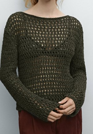 Kobieta ubrana w ciemnozielony, luźno dziergany sweter na szydełku, nałożony na czarną bluzkę, z delikatnie złączonymi dłońmi z przodu oraz widocznymi bordowymi spodniami.