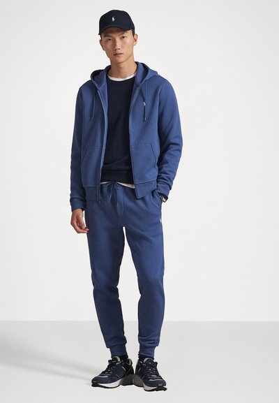 Μπλε φούτερ με κορδόνια, ταιριαστά παντελόνια φόρμας, επικαλυμμένα με ναυτικό μπλουζάκι, και ναυτική καπέλο με λογότυπο; το ύφασμα φαίνεται μαλακό και υφασμένο.