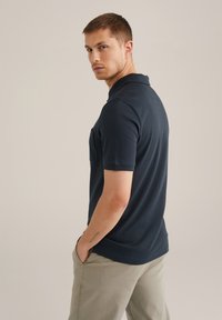 Polo de color azul marino con cuello y mangas cortas, que incluye un bolsillo en el pecho. El modelo lo combinó con pantalones de un color claro, posando en ángulo.